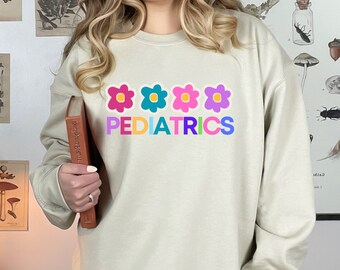 May include: A light beige sweatshirt with a graphic of four colorful flowers and the word "PEDIATRICS" in rainbow colors.