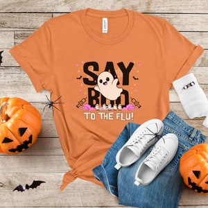 May include: Orange t-shirt with a ghost graphic and the text "Say Boo to the Flu!"  The ghost is wearing a pink bow and has a happy expression. The shirt is laid out on a wooden surface with jeans, white sneakers, and a pumpkin.