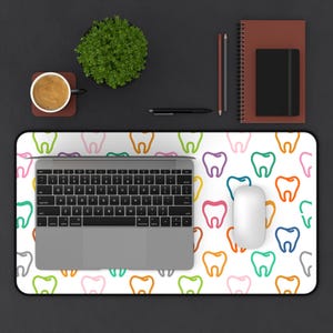 May include: A white desk mat with a colorful tooth pattern, a laptop, and a white mouse. The desk setup includes a cup of coffee, a small plant, a pen, pencils, and a notebook.
