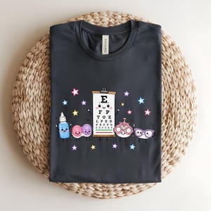 May include: A dark grey t-shirt featuring a graphic print of a smiling eye chart, a blue eye drop bottle, a contact lens case, a phoropter, and purple glasses, all surrounded by small colourful stars against a solid dark grey background.