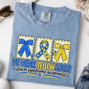May include: A light blue t-shirt featuring a graphic with three scalloped-edge rectangles containing a blue bow, a blue and yellow awareness ribbon, and a yellow bow. The text reads WE WEAR YELLOW & BLUE down syndrome awareness.