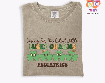 May include: A light brown t-shirt with the text "Caring For The Cutest Little Lucky Charms Pediatrics" and a row of four green shamrock graphics with faces.