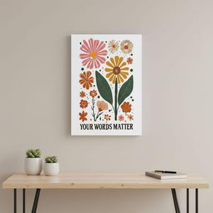May include: A white rectangular art print featuring a floral design with various flowers in shades of pink, orange, and yellow, with green leaves. The text "YOUR WORDS MATTER" is at the bottom in black. The artwork is displayed on a light wooden surface.