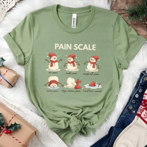 May include: Sage green t-shirt featuring a graphic of snowmen illustrating a "Pain Scale" from "no pain" to "worst pain possible." The snowmen wear red hats and scarves. The shirt has a crew neck and short sleeves.