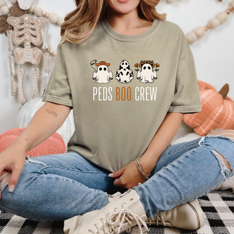 Puede incluir: Una camiseta marr&oacute;n claro con un gr&aacute;fico de tres fantasmas con diferentes disfraces. El texto "PEDS BOO CREW" est&aacute; impreso debajo de los fantasmas.