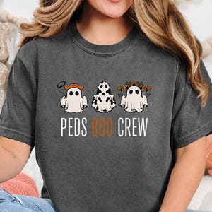 Puede incluir: Una camiseta gris oscuro con un gr&aacute;fico de tres fantasmas con diferentes disfraces. Los fantasmas llevan un sombrero de vaquero, un pa&ntilde;uelo de vaca y una corona de flores. El texto "PEDS BOO CREW" est&aacute; impreso debajo de los fantasmas.