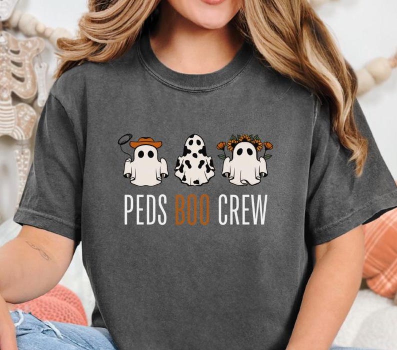Puede incluir: Una camiseta gris oscuro con un gr&aacute;fico de tres fantasmas con diferentes disfraces. Los fantasmas llevan un sombrero de vaquero, un pa&ntilde;uelo de vaca y una corona de flores. El texto "PEDS BOO CREW" est&aacute; impreso debajo de los fantasmas.