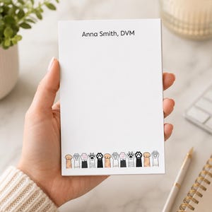 May include: A rectangular white notepad featuring the printed text Anna Smith, DVM at the top. The bottom edge of the notepad is decorated with a horizontal row of various cat paw illustrations in different colors including tan, black, and white.
