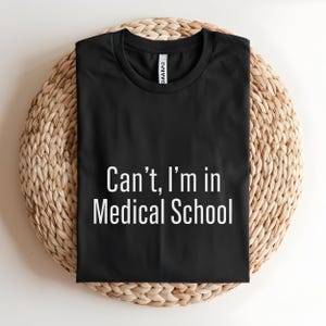 May include: Black t-shirt with the white text "Can't, I'm in Medical School." The shirt is displayed on a round, woven, light brown surface. The shirt has a crew neck and short sleeves.