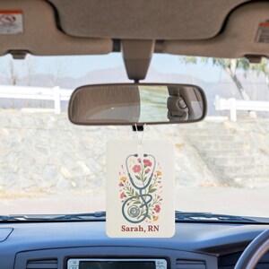 May include: A rectangular white car air freshener hanging from a rearview mirror. The design features a blue stethoscope intertwined with colorful pink, yellow, and green floral illustrations. The text "Sarah, RN" is printed in red at the bottom.