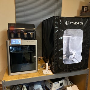 May include: A silver 3D printer with a black top and a clear plastic enclosure. The printer is sitting on a gray metal shelf. A black tent with the text "COMGROW" is on the shelf above the printer.
