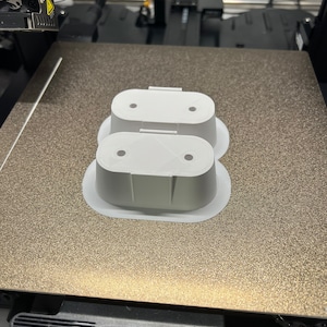 May include: Two white 3D printed objects with rounded edges and two holes on top. The objects are on a textured gray surface.