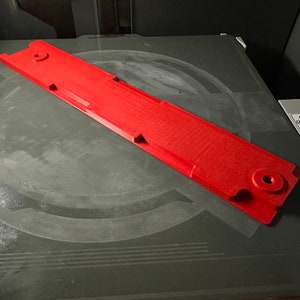 May include: A red 3D printed object with a rectangular shape and several holes. The object is sitting on a gray surface.