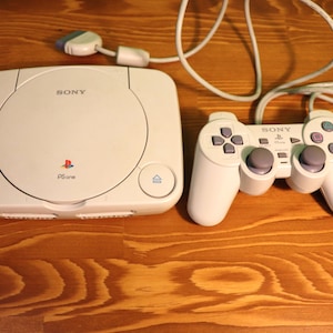Può includere: Un set di console e controller per videogiochi PS one bianchi. La console presenta il logo Sony e la scritta "PS one". Il controller ha un D-pad, pulsanti e due levette analogiche. Il set è su una superficie di legno.