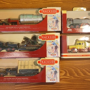 Puede incluir: Cinco camiones de juguete de metal fundido a presión, cada uno con un remolque, en cajas. Los camiones son rojos, negros, amarillos, grises y negros. Las cajas son rojas y blancas con las palabras "Trackside Limited Edition" y "Days Gone" impresas en ellas. Las cajas están sobre una superficie de madera.