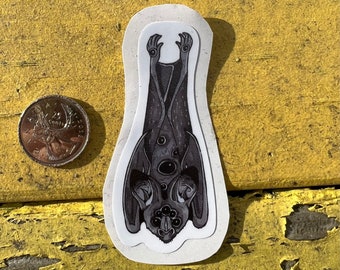 Könnte beinhalten: Ein schwarz-weißer Aufkleber mit einer Fledermaus mit mehreren Augen, die kopfüber hängt. Der Aufkleber befindet sich auf einer gelben Holzoberfläche.