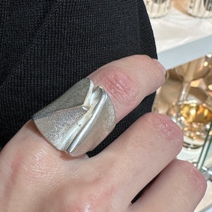 Könnte beinhalten: Ein breiter Silberring mit einer texturierten, matten Oberfläche und einer markanten vertikalen Falte oder Rille in der Mitte. Das Band weist ein geschwungenes, skulpturales Design auf, das sich um den Finger legt und eine geometrische Form bildet.