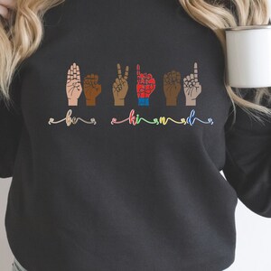 May include: Black sweatshirt with the word "be" and "kind" spelled out in sign language. The hands are different skin tones.