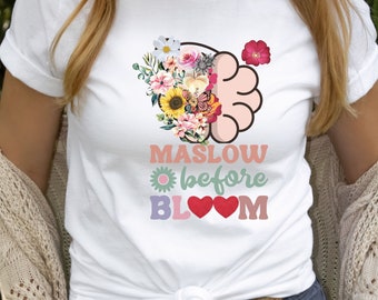 May include: White t-shirt with a floral design and the text "MASLOW before BLOOM" in colorful letters. The floral design includes a sunflower, butterflies, and other flowers.