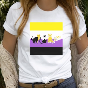 May include: White t-shirt with a graphic of four cats sitting in a row on a yellow, purple, and black striped background. The cats are black, white, and orange.