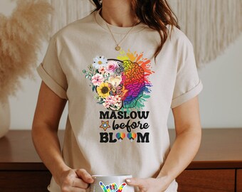 May include: A beige t-shirt with a colorful brain graphic and the text "Maslow before Bloom". The brain graphic is surrounded by flowers and butterflies. The shirt is being worn by a person holding a white mug with a rainbow puzzle piece hand sign.