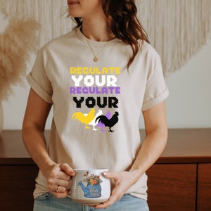 May include: A beige t-shirt with the text "Regulate Your Regulate Your" in pixelated font. Three stylized chickens are below the text, one yellow, one white, and one black. The chickens are standing next to each other.