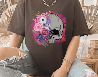 May include: A brown t-shirt with a floral skull graphic. The skull is surrounded by pink, purple, and white flowers. The flowers are painted in a watercolor style.