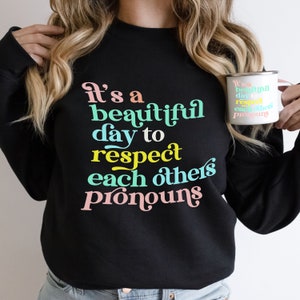 May include: A black sweatshirt with the text "It's a beautiful day to respect each others pronouns" in colorful letters. The text is in a retro font style.