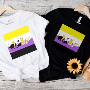 May include: Two t-shirts with a graphic of a non-binary pride flag with cats. The flag is yellow, purple, and black. The t-shirts are white and black.