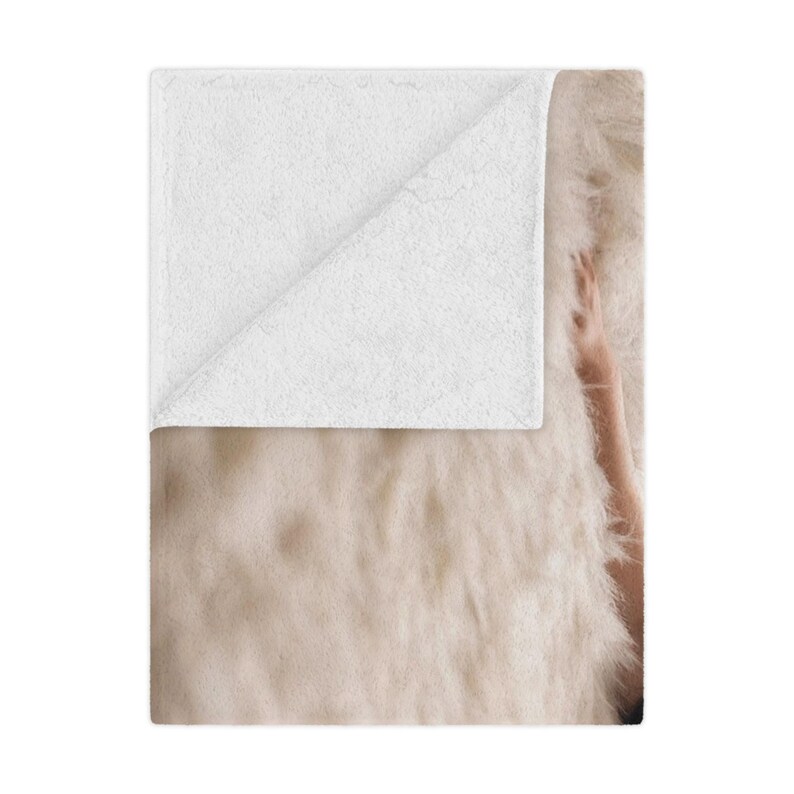 May include: A soft, cream-colored throw blanket with a white, folded corner. The blanket has a fluffy, textured appearance. A person's arm is visible on the right side of the blanket.