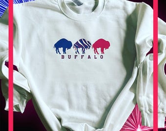 Puede incluir: Sudadera blanca de manga larga con un gráfico de tres siluetas de búfalos. El búfalo del medio está estampado con rayas y los búfalos de la izquierda y la derecha son de color azul y rojo sólido. La palabra "BUFFALO" está impresa debajo de las siluetas de búfalos.