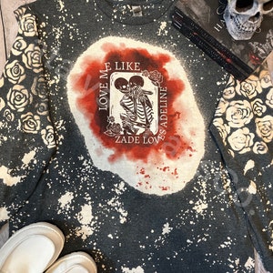 Puede incluir: Una camiseta de manga larga gris oscuro con un patrón de salpicaduras blanqueadas y un estampado gráfico de dos esqueletos abrazados. El texto "LOVE ME LIKE ZADE LOVES ADELINE" está rodeado de acentos rojos y rosas.
