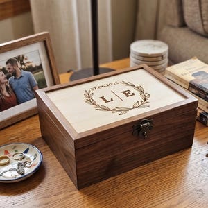 May include: A wooden keepsake box with a light wood lid and dark wood frame. The lid is engraved with the date "27.06.2025", initials "L | E", and a laurel wreath. The box is closed with a metal clasp.