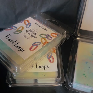 Op de afbeelding: Doorzichtige plastic containers met geurwasmelts. De labels luiden "Froot Loops" en "Candle Delight". De wasmelts zijn pastelkleurig. De achtergrond is een donkere, effen kleur.