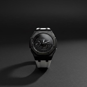 May include: A black and white G-Shock watch with a digital display showing the time. The watch has a black face with white hands and markers, and a white and black strap. The watch is displayed on a dark surface.