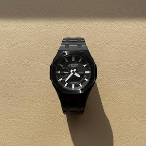 May include: A black G-Shock watch with a black metal strap. The watch face is black with white hands and markers. The watch is displayed on a light brown surface.