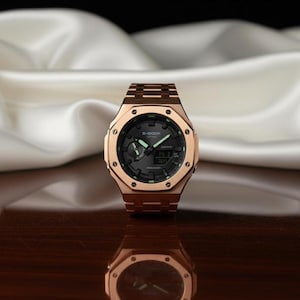 May include: A rose gold-toned watch with a black face and a matching rose gold band. The watch has a digital display and luminous hands. The watch is displayed on a reflective surface.