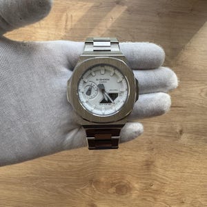 May include: A stainless steel G-Shock Casio watch with a white face and a silver strap. The watch features a digital display and analogue hands. The watch is held by a gloved hand.