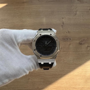 May include: A silver wristwatch with a black face and black strap. The watch has a square face with a circular dial. The watch is held by a gloved hand. The watch face displays the time and date.
