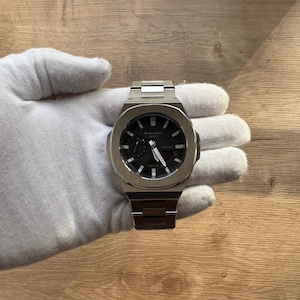 May include: A silver G-Shock watch with a black face and silver hands. The watch features a rectangular case and a stainless steel strap. The watch is held by a gloved hand.