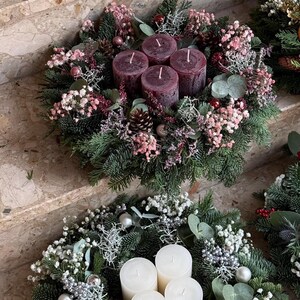 Peut inclure: Deux couronnes de No&euml;l avec des bougies. La couronne sup&eacute;rieure a quatre bougies bordeaux entour&eacute;es de verdure, de fleurs roses et de pommes de pin. La couronne inf&eacute;rieure a quatre bougies blanches, des ornements argent&eacute;s et des fleurs blanches.