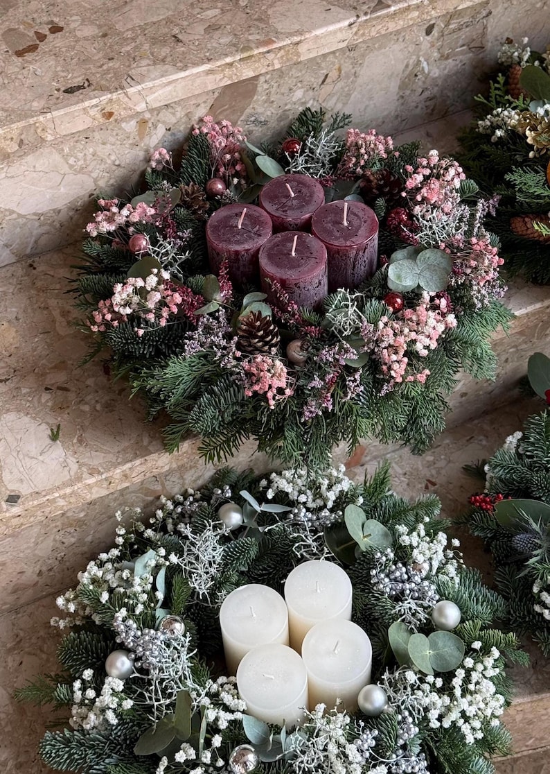 Peut inclure: Deux couronnes de No&euml;l avec des bougies. La couronne sup&eacute;rieure a quatre bougies bordeaux entour&eacute;es de verdure, de fleurs roses et de pommes de pin. La couronne inf&eacute;rieure a quatre bougies blanches, des ornements argent&eacute;s et des fleurs blanches.