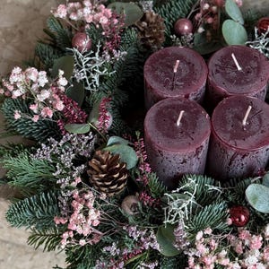 Peut inclure: Une couronne de No&euml;l festive avec quatre bougies bordeaux. La couronne est orn&eacute;e de branches de conif&egrave;res, de fleurs roses, de pommes de pin et de d&eacute;corations. Les bougies ont des m&egrave;ches blanches, cr&eacute;ant une ambiance chaleureuse.