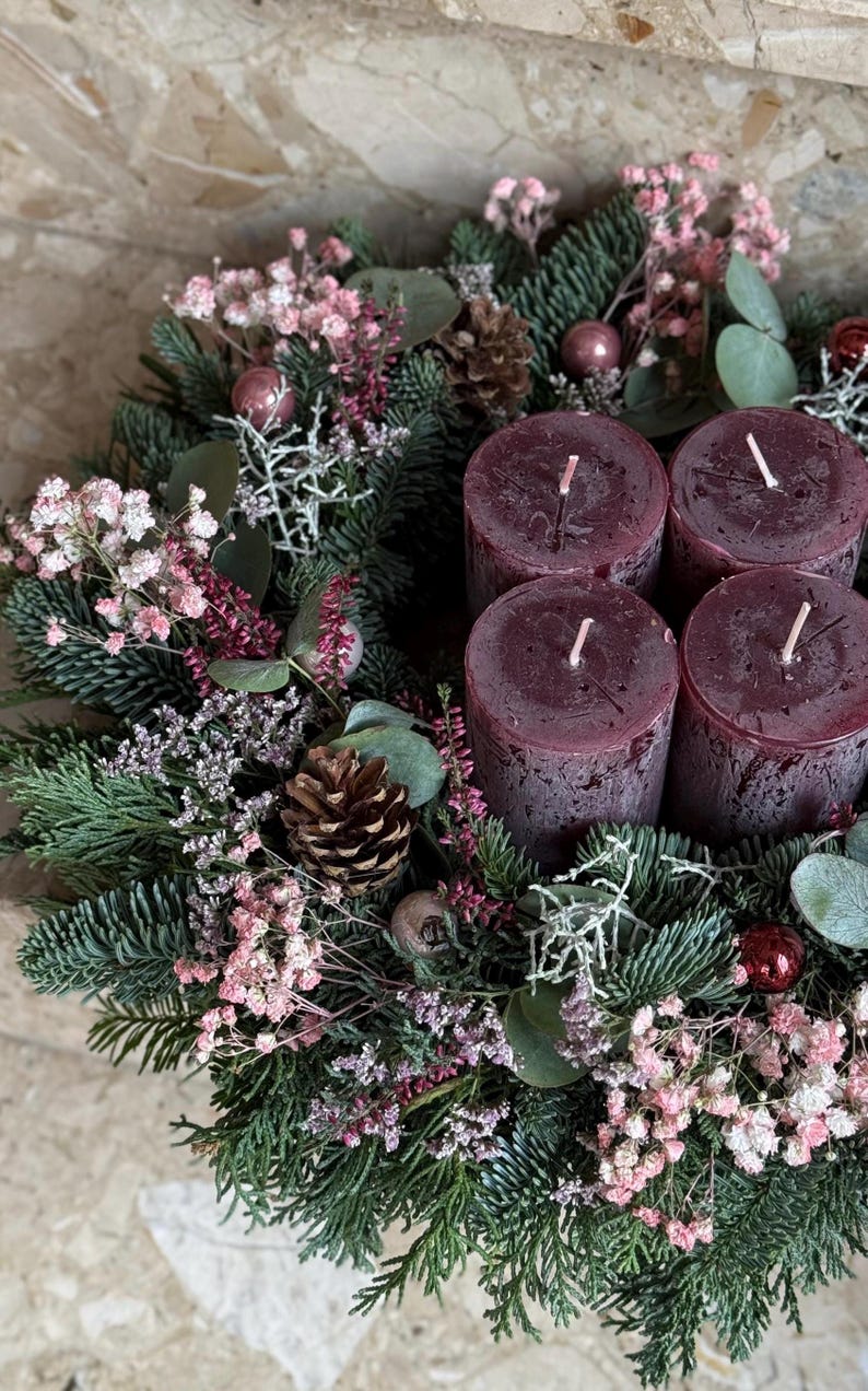 Peut inclure: Une couronne de No&euml;l festive avec quatre bougies bordeaux. La couronne est orn&eacute;e de branches de conif&egrave;res, de fleurs roses, de pommes de pin et de d&eacute;corations. Les bougies ont des m&egrave;ches blanches, cr&eacute;ant une ambiance chaleureuse.