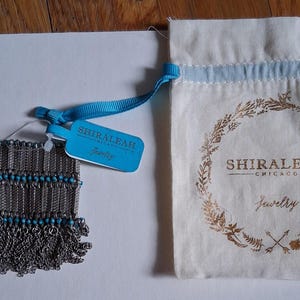 Op de afbeelding: Een zilverkleurige armband met turquoise kralen en franjes, naast een trekkoordtas met de gouden tekst "SHIRALEAH CHICAGO Jewelry". Een blauw label met de merknaam is aan de armband bevestigd.