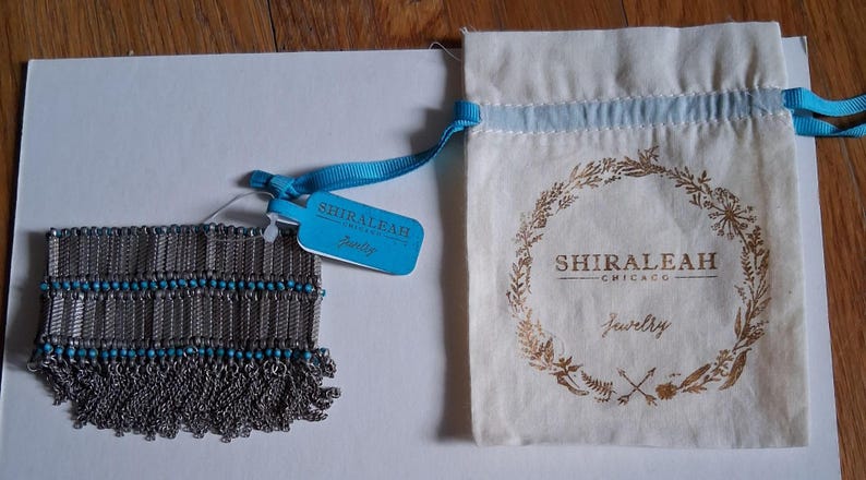 Op de afbeelding: Een zilverkleurige armband met turquoise kralen en franjes, naast een trekkoordtas met de gouden tekst "SHIRALEAH CHICAGO Jewelry". Een blauw label met de merknaam is aan de armband bevestigd.
