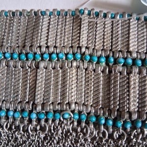 Op de afbeelding: Close-up van een zilverkleurige metalen armband met turquoise kralen. De armband heeft een mali&euml;nkolderontwerp met rechthoekige schakels en kleine, ronde kralen. De schakels hebben een getextureerd, gedraaid patroon.