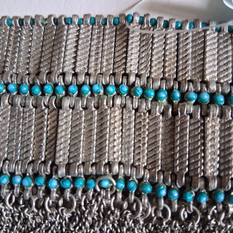 Op de afbeelding: Close-up van een zilverkleurige metalen armband met turquoise kralen. De armband heeft een mali&euml;nkolderontwerp met rechthoekige schakels en kleine, ronde kralen. De schakels hebben een getextureerd, gedraaid patroon.