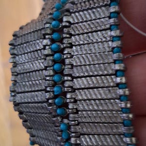 Op de afbeelding: Close-up van een zilverkleurige armband met een geweven ontwerp. De armband heeft rijen rechthoekige schakels, elk met een textuurpatroon. Kleine, ronde, turquoise gekleurde kralen zijn langs de randen verspreid.