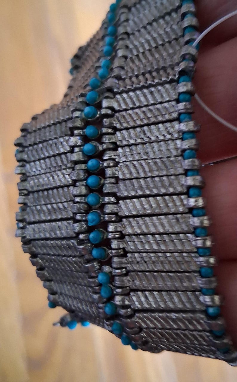 Op de afbeelding: Close-up van een zilverkleurige armband met een geweven ontwerp. De armband heeft rijen rechthoekige schakels, elk met een textuurpatroon. Kleine, ronde, turquoise gekleurde kralen zijn langs de randen verspreid.